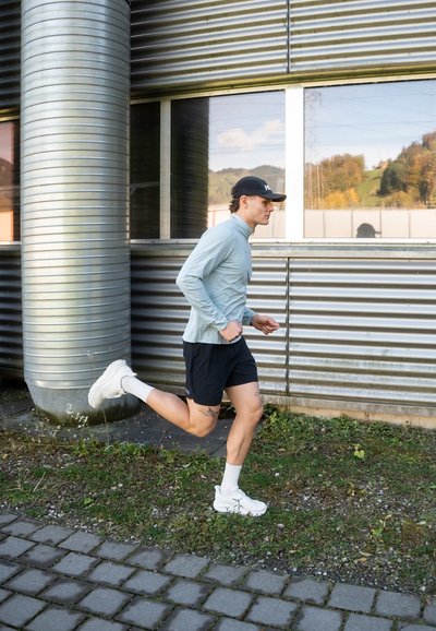 Hombre con ropa deportiva corriendo junto a un edificio de estilo industrial de metal con una gran ventana que refleja un paisaje verde.