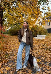 Brun skinnjakke, kremfarget genser, lyseblå vide bukser, hvite joggesko; bærer en brun veske med et lyseblått skjerf. Høstløv i bakgrunnen.