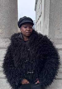 Personne portant un manteau noir duveteux avec des fermoirs métalliques et une casquette gavroche noire, debout entre de grandes colonnes de pierre en extérieur.