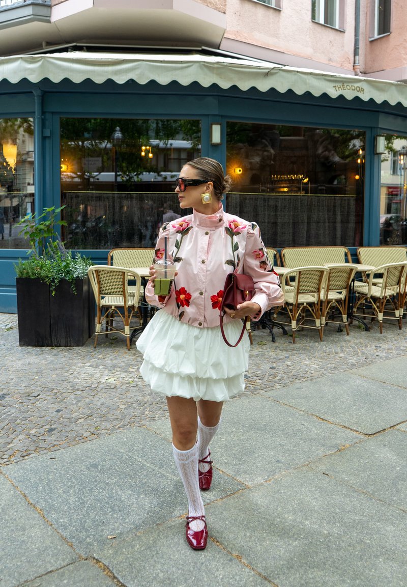 Femme en veste fleurie et jupe blanche à volants marchant à l'extérieur, tenant une boisson et un sac, avec des places assises de café en plein air derrière elle.