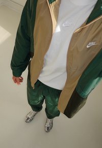 Nike roheline ja beež zip-up jakk, valge särk, rohelised spordipüksid ja hõbedased tennisetossud, esitlevad läikivat, sujuvat tekstuuri ja sportlikku disaini.