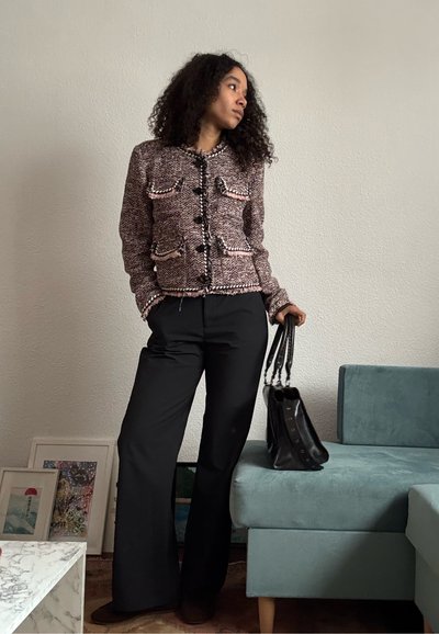Mujer con chaqueta texturizada rosa y negra, pantalones negros de pierna ancha, sosteniendo un bolso negro, de pie cerca de un sofá color azul verdoso y una obra de arte enmarcada.