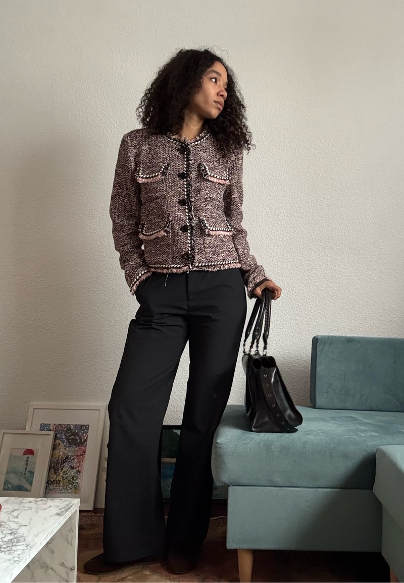 Femme portant une veste texturée rose et noire, un pantalon large noir, tenant un sac à main noir, debout près d'un canapé bleu sarcelle et d'une œuvre encadrée.
