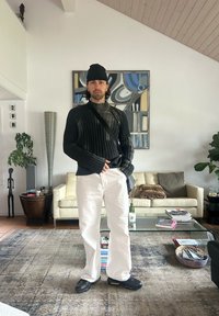 Czarny prążkowany sweter z długimi rękawami, białe spodnie o prostych nogawkach oraz czarne sportowe buty. Nowoczesne wnętrze z zieloną rośliną i dziełem sztuki w tle.