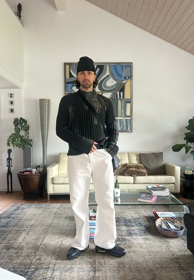 Suéter negro acanalado de manga larga, pantalones blancos de pierna recta y zapatillas negras. Fondo de interior moderno con una planta verde y una obra de arte.