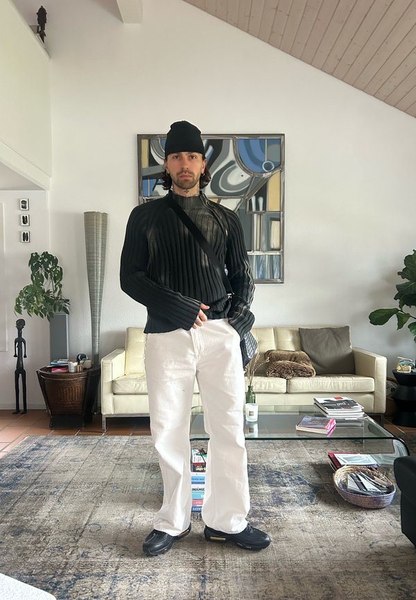 Black ribbed jumper with long sleeves, white straight-leg trousers, and black trainers. Modern interior backdrop with a green plant and artwork.