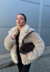 Kremasto pobeljeno, puhasto umetno krzno z ovratnikom šala; nošeno čez temno majico, združeno z okroglo rjavo usnjeno torbico na teksturirani površini.