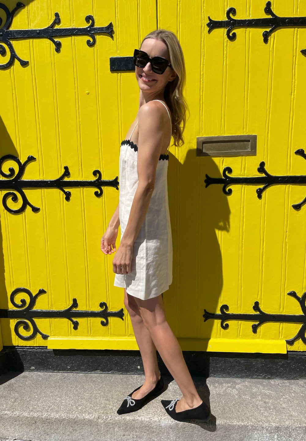 Witte linnen jurk met zwarte golvende randen, gecombineerd met zwarte flats met decoratieve strikken. Heldergele houten deur als achtergrond.