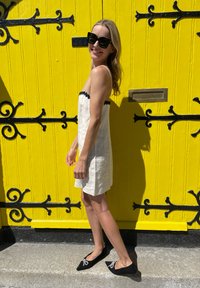 Witte linnen jurk met zwarte golvende randen, gecombineerd met zwarte flats met decoratieve strikken. Heldergele houten deur als achtergrond.