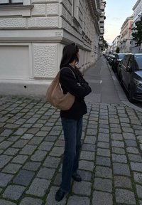 Osoba z długimi ciemnymi włosami, ubrana w czarną marynarkę i dżinsy, stoi na kostkowej ulicy, patrząc wzdłuż szeregu zaparkowanych samochodów i budynków.