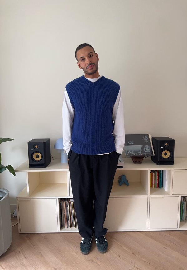 Uomo in piedi all'interno indossando un maglione smanicato blu sopra una camicia bianca a maniche lunghe, pantaloni neri e scarpe da ginnastica, con altoparlanti e dischi dietro di lui.