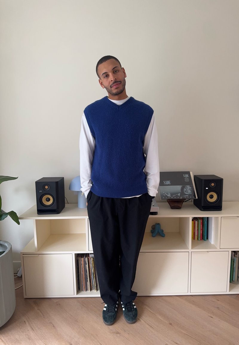 Man som står inomhus klädd i en blå ärmlös tröja över en vit långärmad skjorta, svarta byxor och sneakers, med högtalare och skivor bakom sig.
