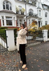 Femme en manteau en fourrure beige et lunettes de soleil se tenant sur une rue pavée devant des maisons de ville blanches avec des feuilles d'automne au sol.