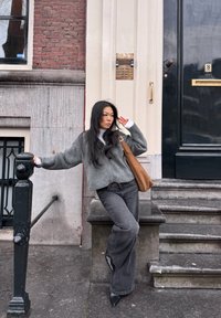 Jeune femme en pull gris et jean, appuyée sur une rambarde noire à l'extérieur d'un bâtiment en brique et pierre avec une porte sombre et des escaliers.