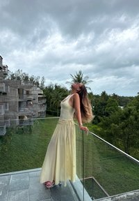 Lang gul kjole med en tætsiddende overdel og en flydende nederdel, kombineret med lyserøde sandaler. Omgivelserne har et glasgelænder og tropisk grønt områder.