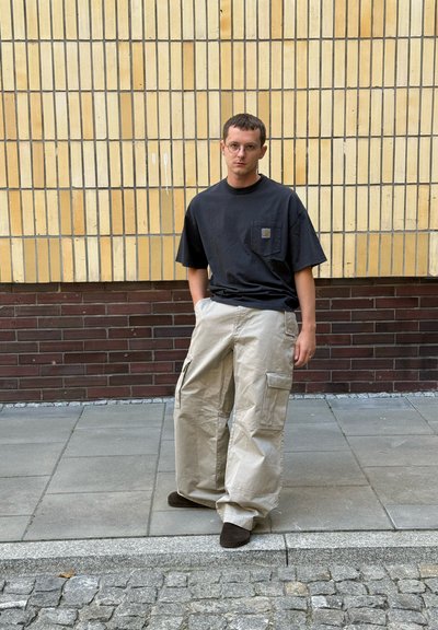 Camiseta negra de manga corta con un bolsillo en el pecho; pantalones cargo beige con múltiples bolsillos; zapatos marrones. Fondo neutro con pared texturizada.