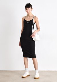 Vestido midi negro ajustado con tirantes finos, que presenta un pequeño logo. Combinado con zapatillas blancas y un pequeño bolso bandolera blanco.