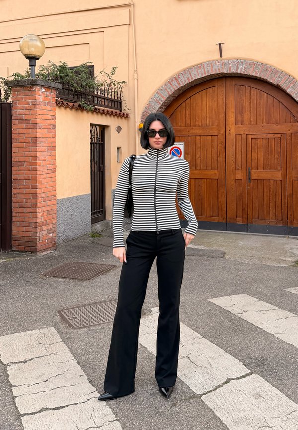 Femme portant des lunettes de soleil noires, un haut rayé zippé, un pantalon noir, debout sur un passage piéton devant une grande porte en bois arquée.