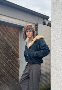 Jeune personne portant une veste en denim doublée de fourrure et un chapeau de trappeur en fourrure, debout près d'une porte en bois et d'une grille en métal, regardant sur le côté.