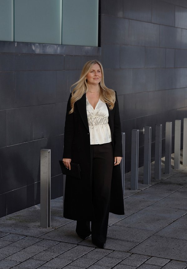 Donna con lunghi capelli biondi che cammina su un marciapiede urbano, indossando un lungo cappotto nero, una camicetta bianca, pantaloni neri e tenendo una pochette nera.