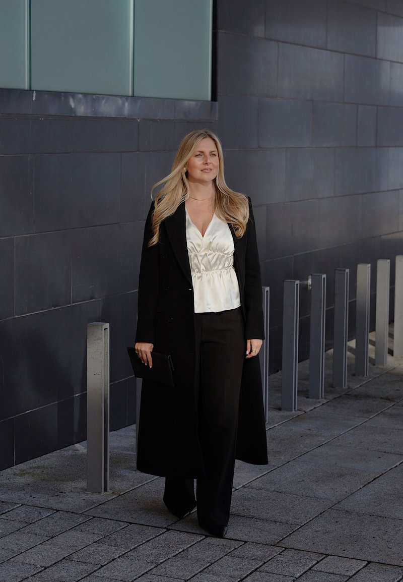 Frau mit langen blonden Haaren, die auf dem städtischen Bürgersteig geht, trägt einen schwarzen langen Mantel, eine weiße Bluse, schwarze Hosen und hält eine schwarze Clutch-Tasche.