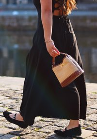 Vævet stråhåndtaske med brune læderdetaljer, der har et rundt top håndtag og et guld logo. Sort maxikjole og flade sorte sko.