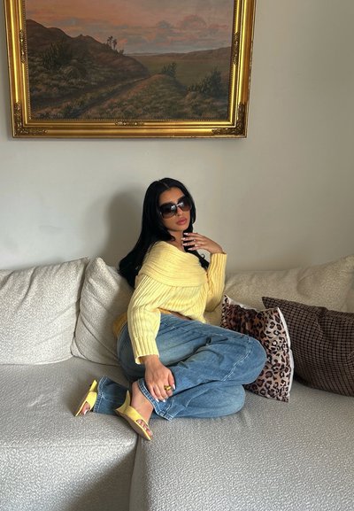 Mujer con suéter amarillo, jeans azules y tacones amarillos sentada en un sofá beige con cojines estampados y un cuadro de paisaje en la pared detrás.