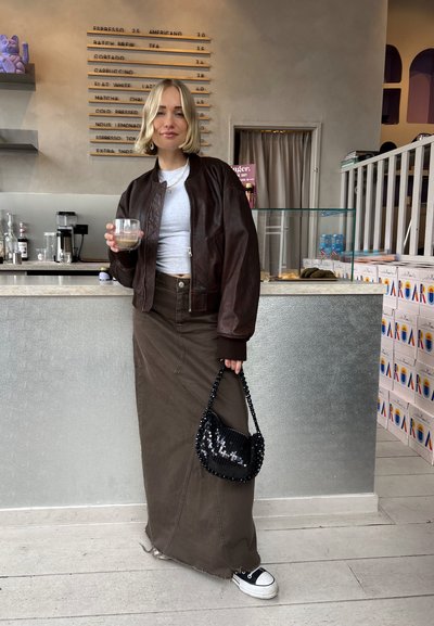Chaqueta bomber de cuero marrón sobre una camiseta gris, falda larga verde oliva, bolso de mano con lentejuelas negras, zapatillas blancas, de pie en una cafetería.