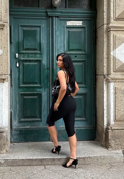 Vestido negro ajustado con tirantes, combinado con zapatos de tacón alto negros y un bolso negro. Puertas de madera verdes al fondo.