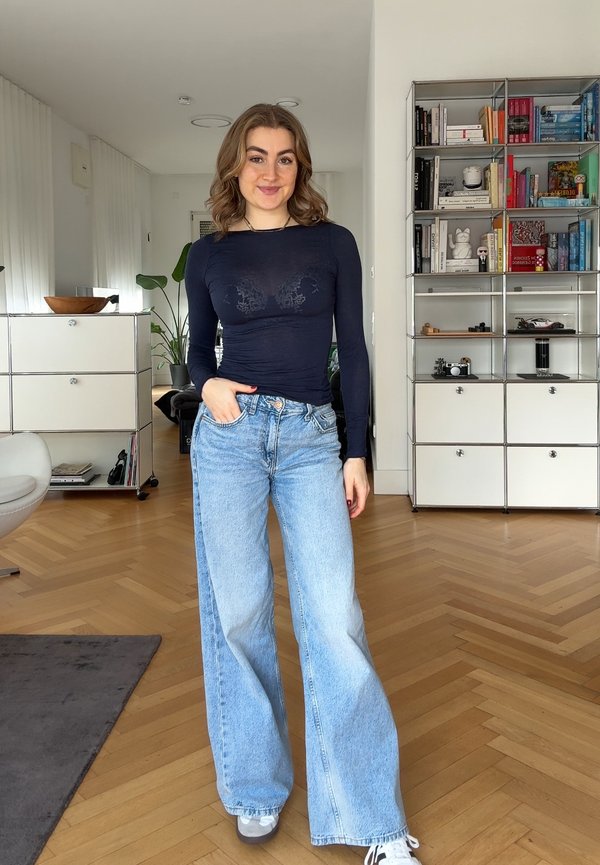 Jeune femme portant un haut ajusté à manches longues de couleur sombre et un jean bleu clair à jambes larges, debout dans un salon moderne avec un sol en bois.