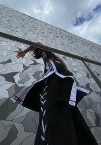 Tenue de sport noire avec des rayures blanches, comprenant une longue jupe avec des détails en lanières et des boutons-pression, devant un mur à motif géométrique.