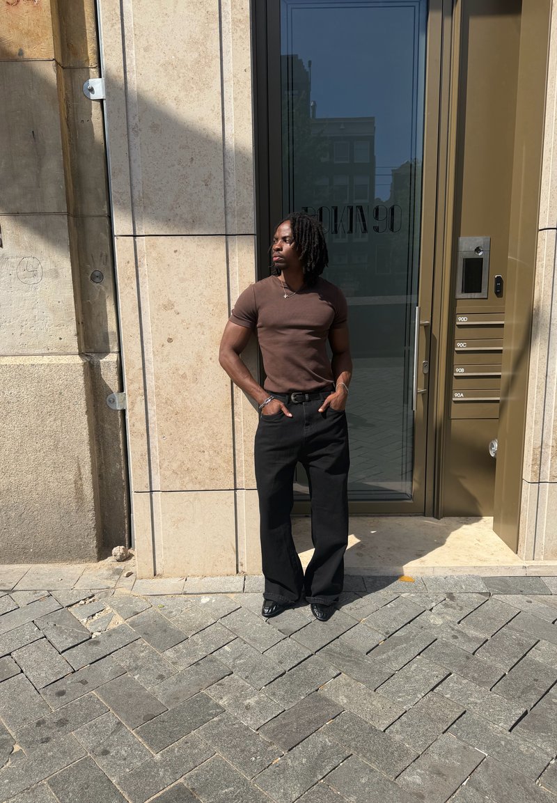Homem com dreadlocks escuros, vestindo uma camisa castanha e calças pretas, está em pé com as mãos nos bolsos à entrada do edifício.