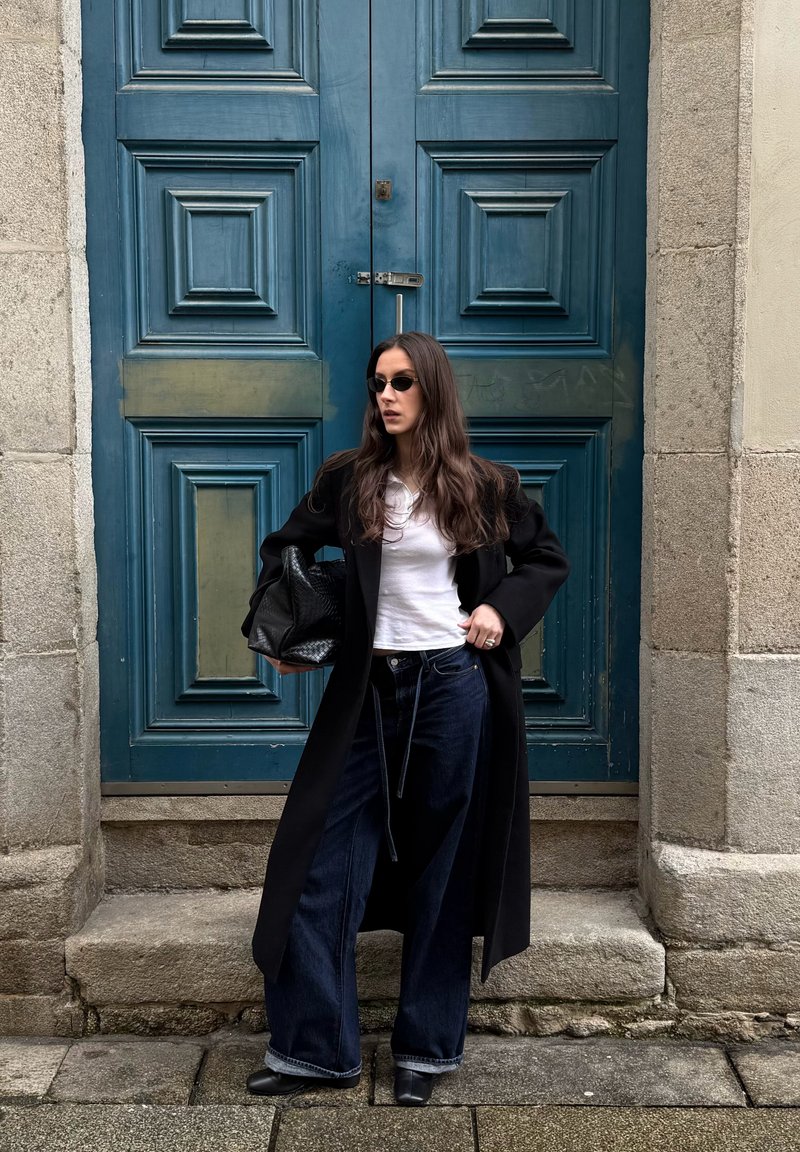 Femme portant un manteau noir, une chemise blanche, un jean large, des lunettes de soleil, tenant un sac à main noir, debout devant une grande porte en bois bleue avec un cadre en pierre.