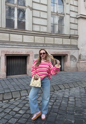 Mujer con suéter de rayas rosas, jeans azul claro y zapatos de plataforma, de pie sobre una calle empedrada sosteniendo un bolso beige, con un edificio histórico de fondo.