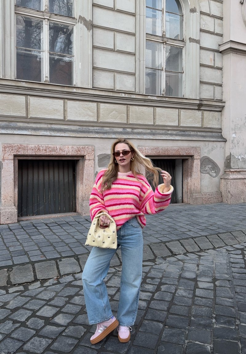Donna con maglione a righe rosa, jeans azzurri e scarpe con plateau, in piedi su una strada acciottolata con una borsa beige in mano, sfondo di edificio storico.