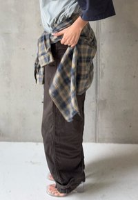 Personne portant un pantalon marron foncé et des talons transparents, tenant une chemise à carreaux bleue et jaune nouée autour de la taille contre un fond uni.