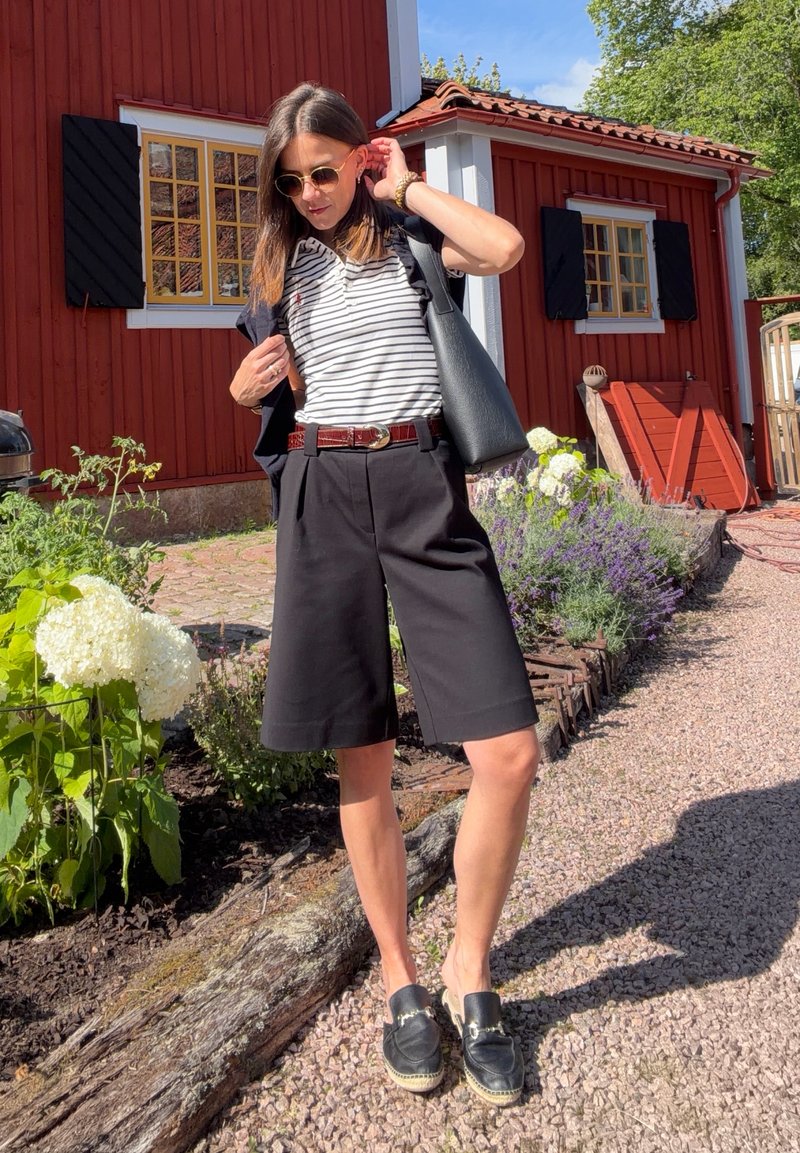 Femme portant des lunettes de soleil, une chemise rayée, un short noir et des chaussures sans lacets, debout sur un chemin de gravier près d'une maison en bois rouge et d'un jardin fleuri.