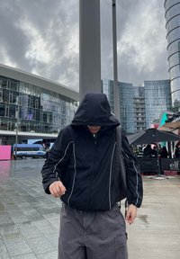 Veste zippée noire avec des accents blancs, capuche relevée, associée à un pantalon gris ample. L'arrière-plan présente des bâtiments en verre et un ciel nuageux.