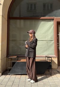 Sort oversized blazer, brune bredde bukser og en mørk baseballkasket. Lyse sneakers. Minimalistisk metal møbel i baggrunden.