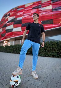 Μαύρο μπλουζάκι με ροζ λογότυπο, ανοιχτό μπλε παντελόνι και μπεζ αθλητικά παπούτσια. Στέκεται σε μια μοτίβο πεζοδρόμιο με μια πολύχρωμη μπάλα ποδοσφαίρου.