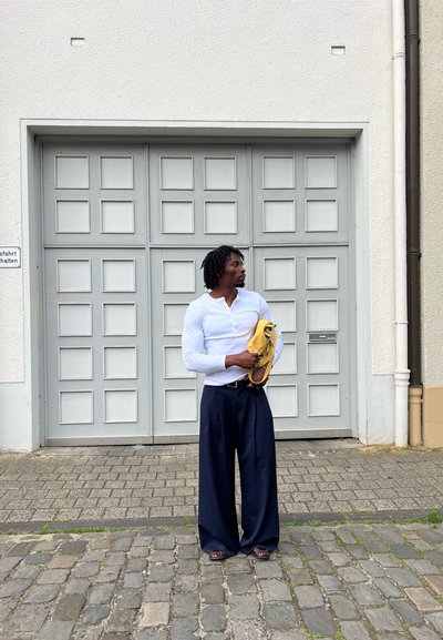 Camisa blanca de manga larga, pantalones anchos azul marino, sosteniendo un bolso amarillo. El fondo presenta una puerta de garage gris con paneles cuadrados.