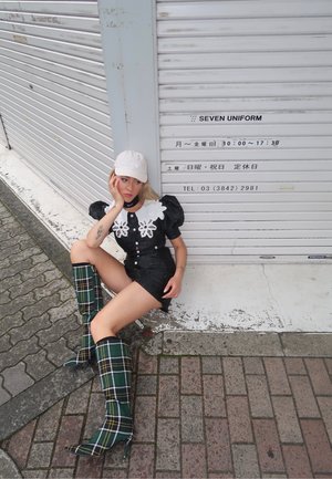 Vestido corto negro con mangas abullonadas y cuello floral blanco, combinado con botas de cuadros verdes hasta la rodilla. Lleva una gorra blanca, sentada contra una pared.