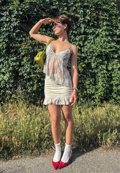 Top de tie-dye pastel, fluido, falda crema ajustada con volantes, calcetines blancos y tacones rojos de punta. La modelo sostiene un bolso verde lima, de pie al aire libre.