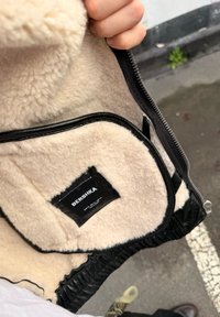 Main tenant une veste noire ouverte avec une doublure en polaire beige et une étiquette de la marque Bershka visible à l'intérieur.