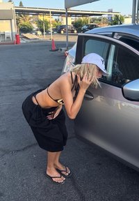 Person iført en sort crop top og oversized bukser, der læner sig ind mod en grå bil. Baseball cap, sorte flip-flops og lyst blond hår synligt.