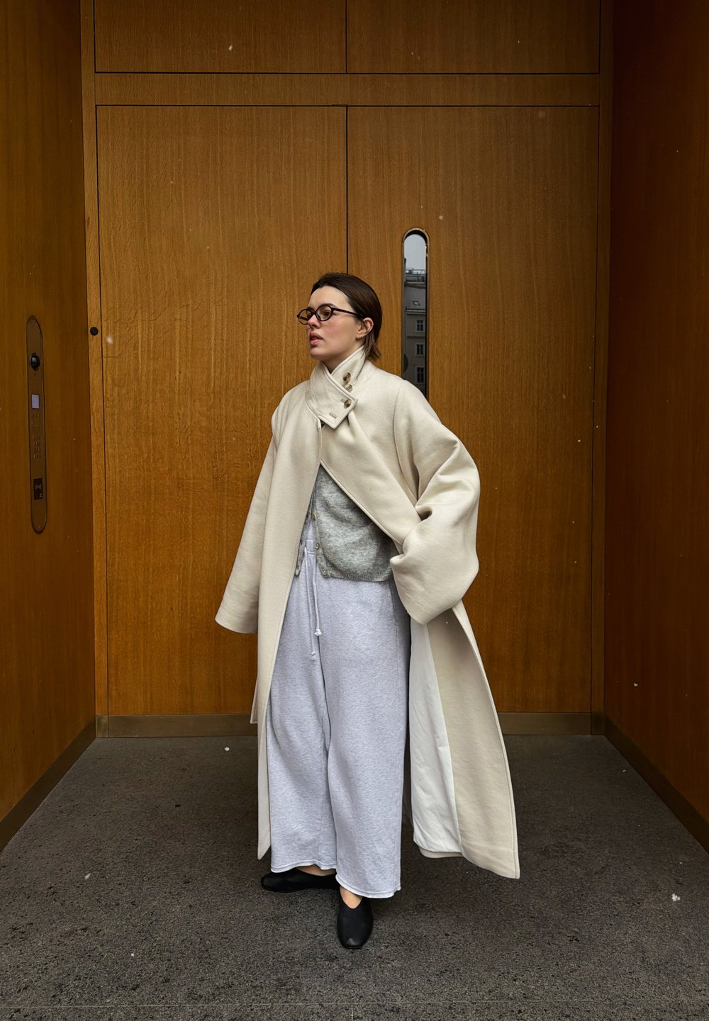 Femme portant un manteau beige surdimensionné, un pull gris, un pantalon gris ample et des chaussures noires, debout devant des portes d'ascenseur en bois.
