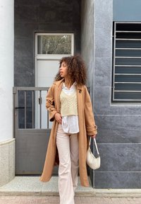 Cappotto lungo color cammello sopra una camicetta bianca e un gilet lavorato a maglia crema. Pantaloni in corduroy chiari e una piccola borsa bianca. Sfondo di una parete tessurizzata.