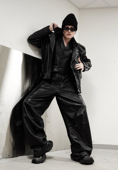Chaqueta de cuero negra sobre un chaleco negro y pantalones de pierna ancha, combinados con zapatillas negras gruesas. Los accesorios incluyen gafas de sol oscuras y un gorro.
