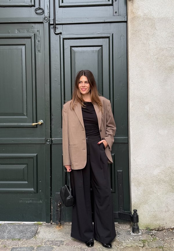 Mujer con cabello largo y castaño está sonriendo frente a una puerta de madera de color verde oscuro, lleva puesto un blazer beige, un top negro, pantalones negros anchos y sostiene un bolso negro.