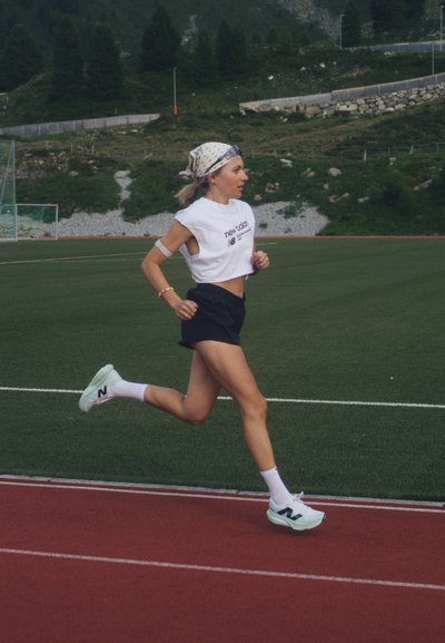 Atleta corriendo en una pista, vistiendo una camiseta corta blanca y pantalones cortos negros. Los zapatos son de color verde claro con un logo distintivo. El fondo presenta vegetación.